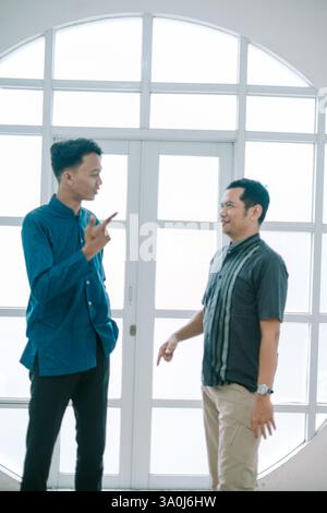 Two asian men are having a discussion near a window in a bright indoor setting. They are gesturing and seem engaged in a lively conversation about a p Stock Photo