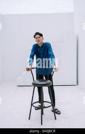 Portrait of a thoughtful young man in casual attire leaning on a chair inside minimalist studio. Clean, bright background with geometric shapes. Suita Stock Photo
