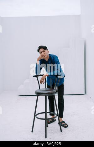 A stylish young man in casual attire leans on a black stool within a bright, minimalist white studio. His thoughtful expression suggests contemplation Stock Photo