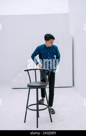 A pensive young man wearing a stylish blue shirt and black pants is leaning on a black bar stool in a minimalist white studio. He exudes a modern and Stock Photo