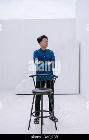A young asian man wearing a blue shirt standing confidently with his arms crossed in a minimalist white studio setting. He is casually leaning against Stock Photo