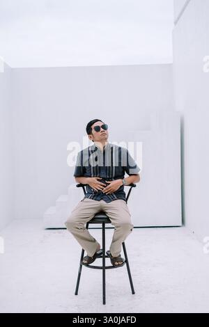 A man wearing sunglasses sits on a chair in a bright white room, gazing upwards with a thoughtful expression. His relaxed posture and casual attire su Stock Photo
