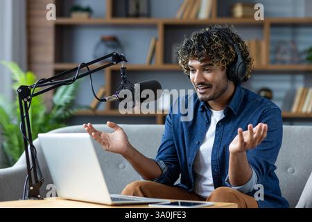 Young man enthusiastically records a podcast or participates in a lively online meeting from his living room, using a microphone and laptop. Stock Photo