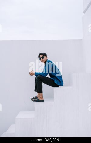 Young stylish man wearing sunglasses sits on white steps with a minimalist architecture background. He is wearing casual clothing and seems thoughtful Stock Photo