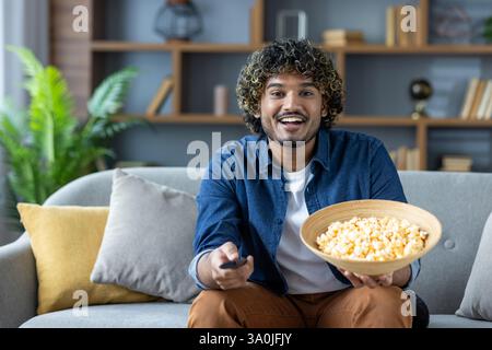 A happy man with curly hair watches television while relaxing on the sofa with a large bowl of popcorn at home. Stock Photo