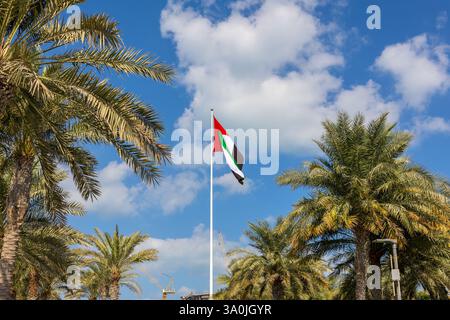 Flag of United Arab Emirates in National Day UAE. Flag is flying against background of blue sky with white clouds and tall green palm leaves. High quality photo Stock Photo