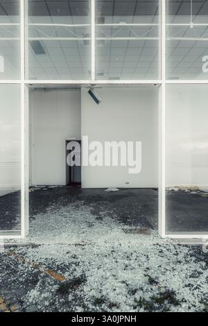 Abandoned commercial building awaiting demolition. Shattered glass covers the ground. Broken storefront windows reveal an empty interior with white wa Stock Photo