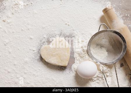 Heart-shaped cookie dough on floured surface with egg, rolling pin, and sieve homemade baking for Valentine's Day or festive culinary concept. Stock Photo