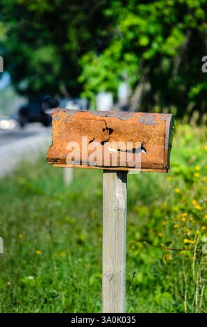 Rusty, dented rural mailbox in front of out of focus trees and a road in the summer. Stock Photo
