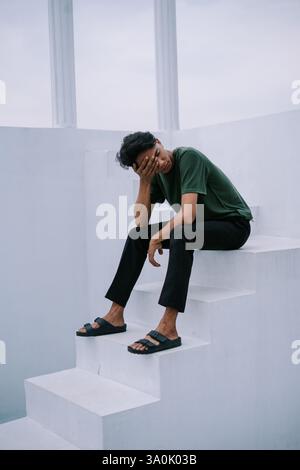 A young man sits on bright white steps, his posture conveying a sense of introspection. He is dressed in casual clothing, and he appears thoughtful am Stock Photo