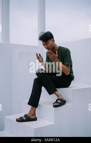 A young man sits on minimalist white stairs, lost in thought. His contemplative expression suggests introspection and reflection on life's journey. Ca Stock Photo