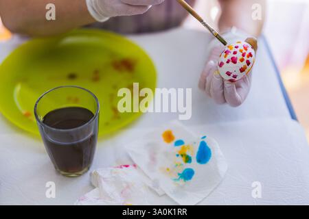 Hand painting Easter egg with red and orange floral pattern using brush on white table with art supplies. Sweden. Stock Photo