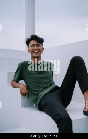 A cheerful young man relaxes on bright white steps, his radiant smile reflecting pure happiness. He's dressed casually, creating an easygoing and invi Stock Photo