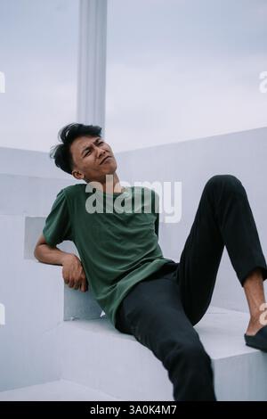 A thoughtful young Indonesian man in a green t-shirt and black pants gazes up at the overcast sky. He's sitting casually on white steps on a rooftop o Stock Photo