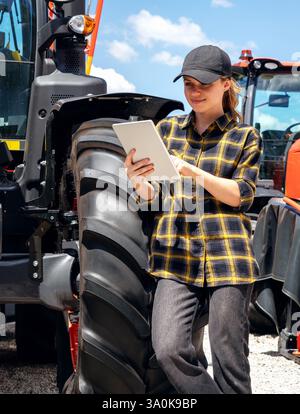 Woman using digital tablet while standing next to agricultural or construction machinery outdoors. Stock Photo