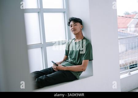 A thoughtful young man is seated indoors, leaning against a bright window frame while holding his smartphone. The modern architecture and casual attir Stock Photo