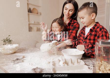 Happy mother and kids baking together, playing with flour in a cozy kitchen, wearing matching plaid shirts, joyful family bonding and messy fun moment Stock Photo