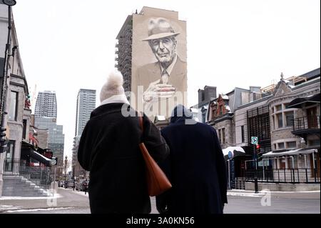 Elder couple, bundled in winter warmth, stands before Leonard Cohen’s iconic mural in downtown Montreal Stock Photo