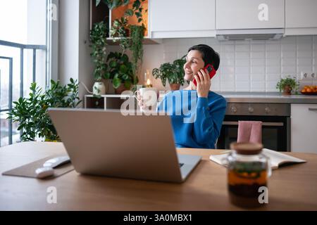 Smiling mature woman enjoy phone call and herbal tea. Relaxed comfortable home job with slow living Stock Photo