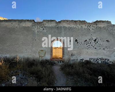 Weathered white stone wall with a red arched brick doorway framing another wall with sunlight Stock Photo