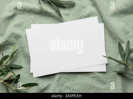 Two blank white cards lie on a textured green fabric adorned with olive branches, creating a stylish mockup for invitations, announcements, or station Stock Photo