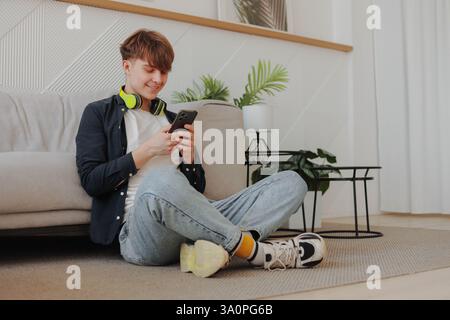 Young smiling man student with yellow headphones using phone at living room. Stock Photo