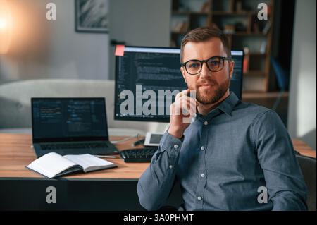 Software developer is sitting and posing. Man in formal clothes is working in the modern office. Using computer. Stock Photo