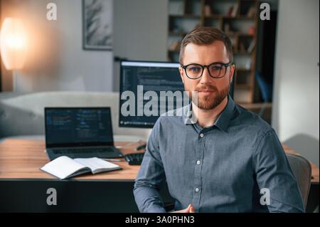 Software developer is sitting and posing. Man in formal clothes is working in the modern office. Using computer. Stock Photo