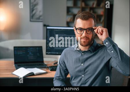 Software developer is sitting and posing. Man in formal clothes is working in the modern office. Using computer. Stock Photo