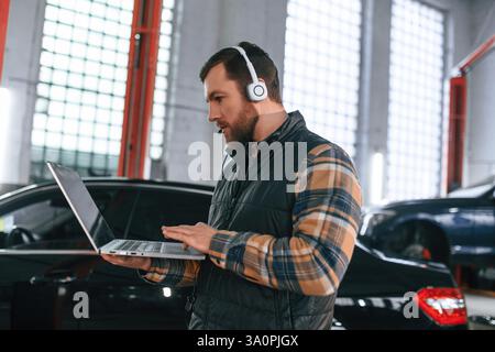 Standing with laptop in hands and in headphones. Man in uniform is working in the auto salon. Stock Photo