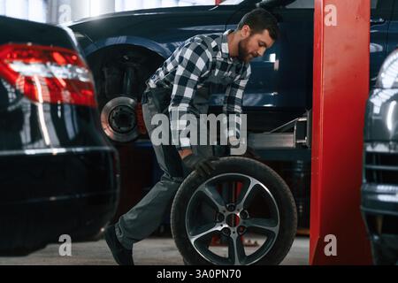 Side view. Moving the tire for the car. Man in uniform is working in the auto salon. Stock Photo
