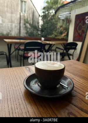 A ceramic cup of coffee with latte art sits on a wooden table in an outdoor cafe with empty tables and chairs in the background. The setting includes Stock Photo