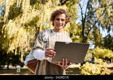 A young man smiles while using his laptop in a vibrant park, surrounded by trees and sunlight. Stock Photo