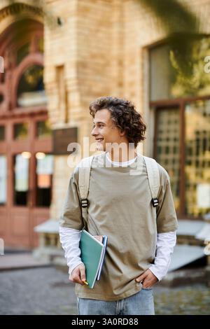 A young man with curly hair walks through a sunlit campus, smiling and holding books. Stock Photo