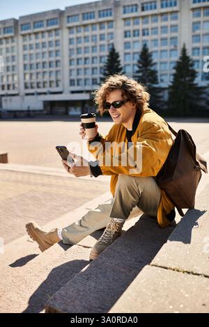 A young stylish man sits on a step, smiling while enjoying his coffee and using his smartphone. Stock Photo