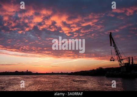 A vibrant sunset over a river, featuring a dramatic sky ablaze with pink and orange clouds. A large industrial crane is silhouetted against the sky, a Stock Photo