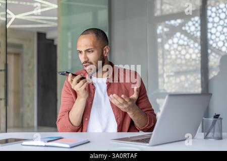 Worried businessman using voice assistant on his smartphone while sitting at his desk in a modern office setting with a laptop. Stock Photo
