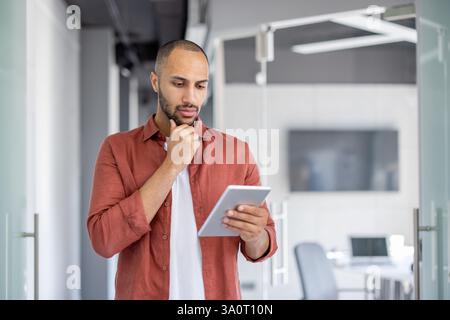 Serious thinking programmer developer standing with tablet computer in hands. Man testing new application software. Confident company employee reading online data. Stock Photo