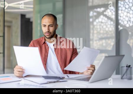 Serious focused financier businessman doing paperwork, man checking documents, papers reports. Office worker inside office thinking working with laptop. Stock Photo