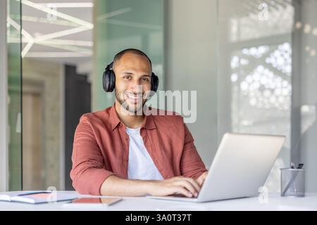 Young successful man working with laptop inside office, using headphones to listen to music at workplace, joyful smiling looking at camera. Stock Photo