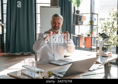 Sitting and smiling. Senior doctor in white coat is at work indoors. Stock Photo
