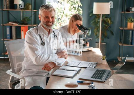 Man is smiling, woman is using microscope. Two clinic workers are indoors together. Stock Photo