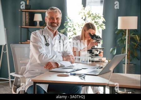 Man is smiling, woman is using microscope. Two clinic workers are indoors together. Stock Photo