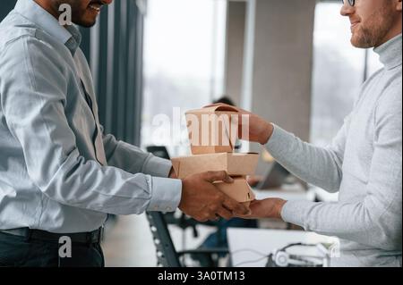 View from the side. Holding paper eco boxes. Two men are working in the modern office together. Stock Photo