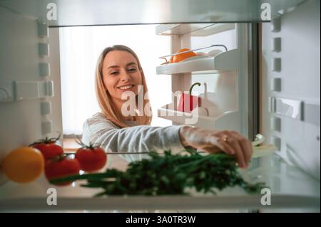 Vegetables is in the fridge. Beautiful young woman is on the kitchen. Stock Photo