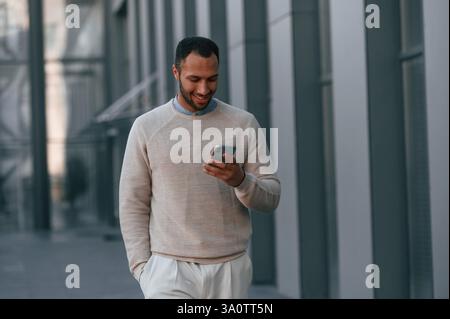 Handsome man is walking, holding smartphone. Outdoors near the business building. Stock Photo
