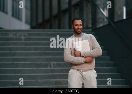 Positive handsome black man is outdoors near the business building holding laptop. Stock Photo
