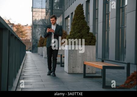 Front view. Using smartphone. Handsome man in formal clothes is outdoor near the business building. Stock Photo
