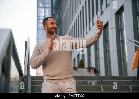 Online conversation, using smartphone. Handsome black man is outdoors near the business building. Stock Photo