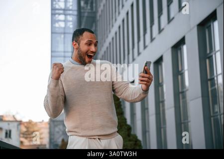Happy guy holding smartphone and celebrating success. Handsome black man is outdoors near the business building. Stock Photo